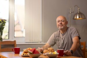 This is a photo of a man sitting at a breakfast table. It is the image at the top of a news page on the Urostomy Association's website. The news article relates to a survey being conducted by Hollister on peristomal skin complications amongst the ostomy community.