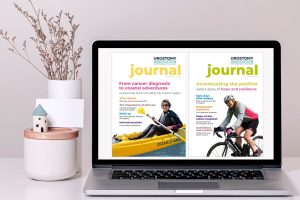 This picture shows a large screen desk top computer, showing the covers of two editions of the Urostomy Association's Journal. To the left of the screen is a vase of dried flowers and a pot.