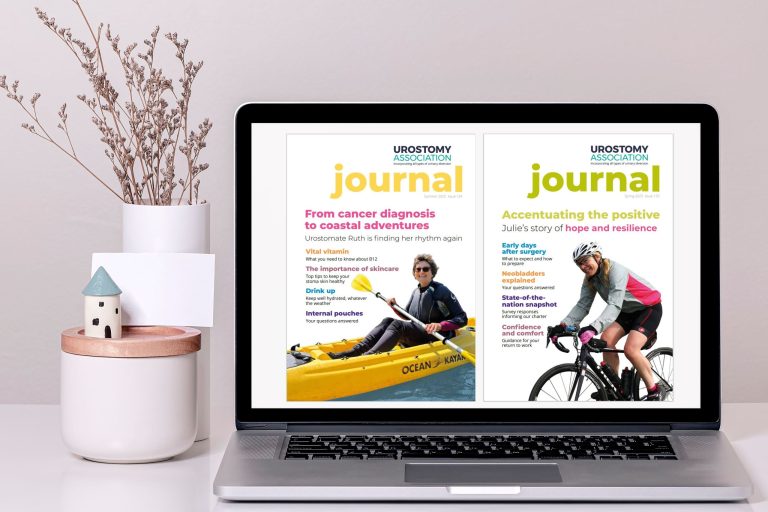 This picture shows a large screen desk top computer, showing the covers of two editions of the Urostomy Association's Journal. To the left of the screen is a vase of dried flowers and a pot.