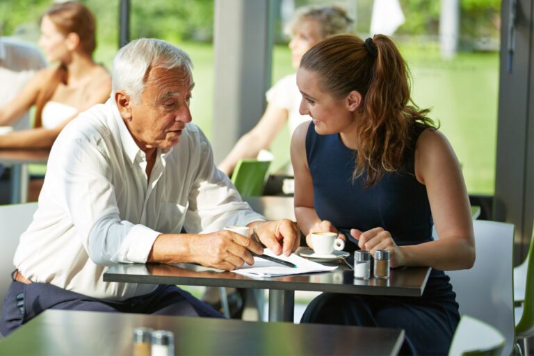 This is a photo of two people sitting in a cafe looking at a notebook. It is the header image for the FAQs page on the Urostomy Association's website.