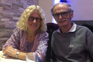 This is a photograph of Allen Rowlinson and his wife, Anneka. They're sitting side by side at a restaurant table, smiling at the camera. It is the header image of a page on the Urostomy Association's website entitled 'Cancer isn't going to beat me'!