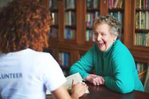 This is a photo of two people sitting in a library talking. The woman in the green top, facing the camera, is laughing. The woman with her back to the camera has the word 'volunteer' written on the back of her t-shirt