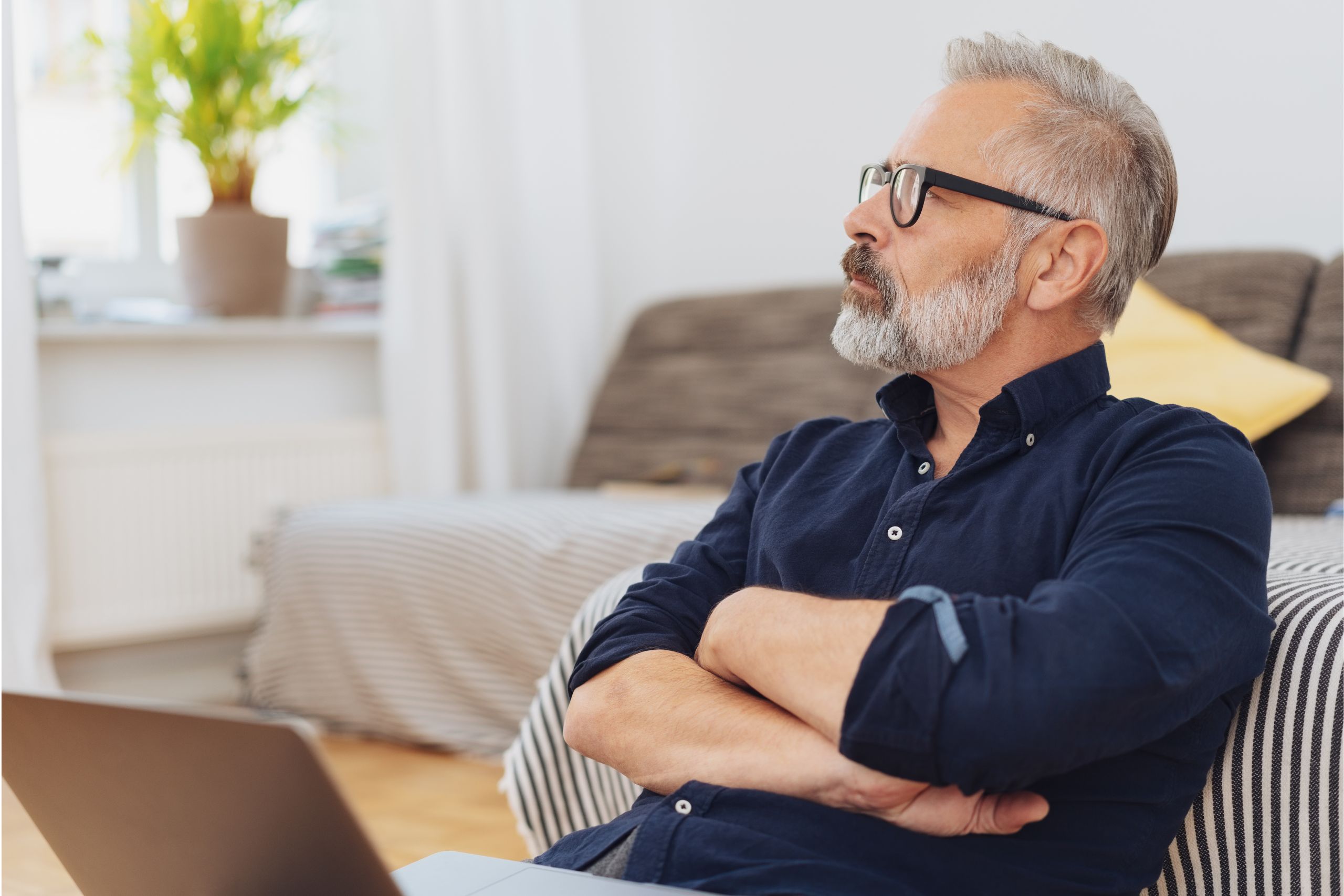 This is a photo of a middle-aged white man, wearing a navy shirt and glasses. He is sitting on a sofa with a laptop on his knee. He is looking into the distance, thoughtful. It is the header image for the 'Preparing for surgery' image on the Urostomy Association's website