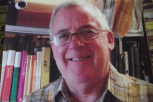 This is a photo of Roger Bowen. He is standing in front of a book case and smiling directly into the camera.