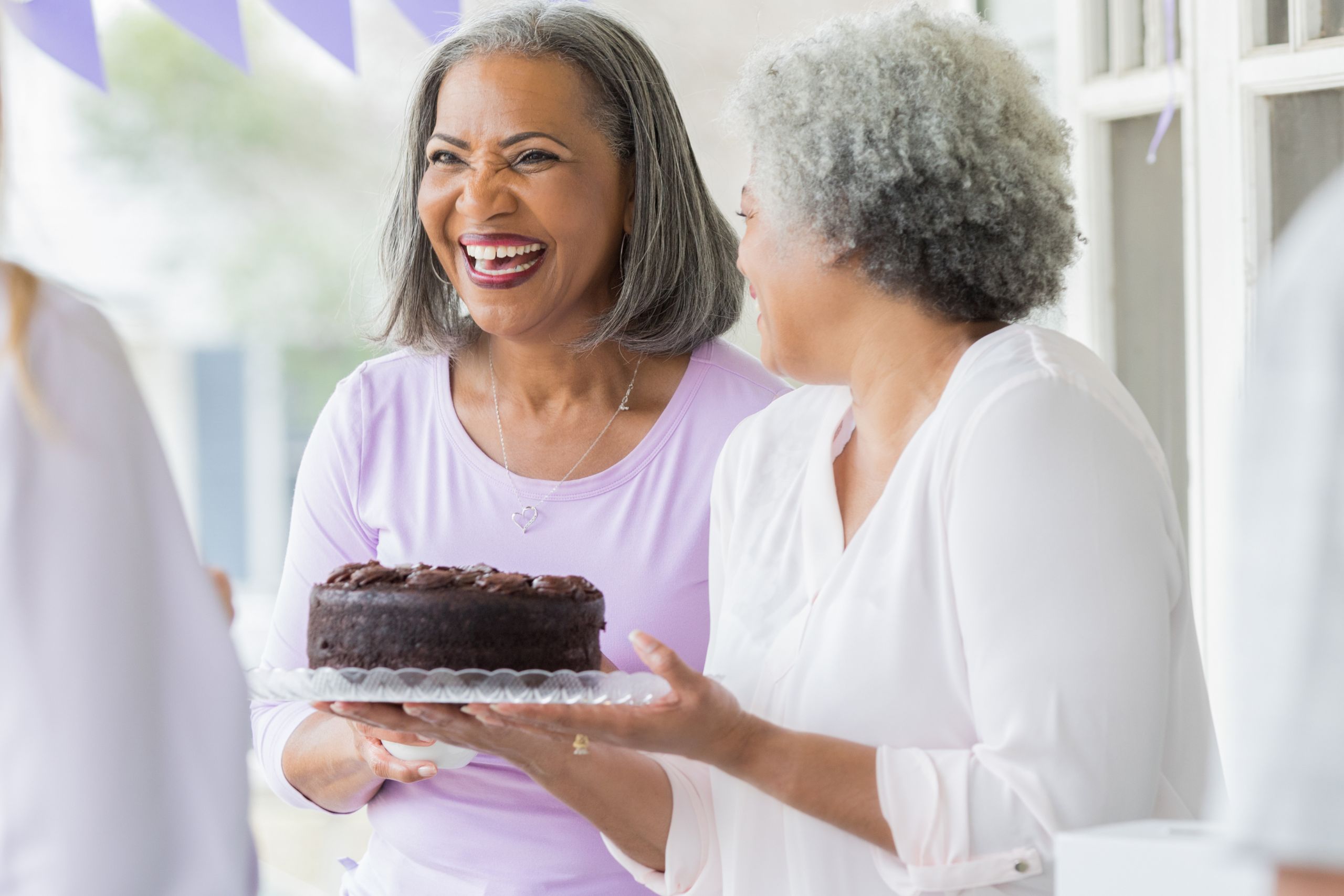 This is a photo of two woman smiling and chatting over a chocolate cake. It is the header image for the Urostomy Association's 'keep it safe and keep it legal' fundraising page