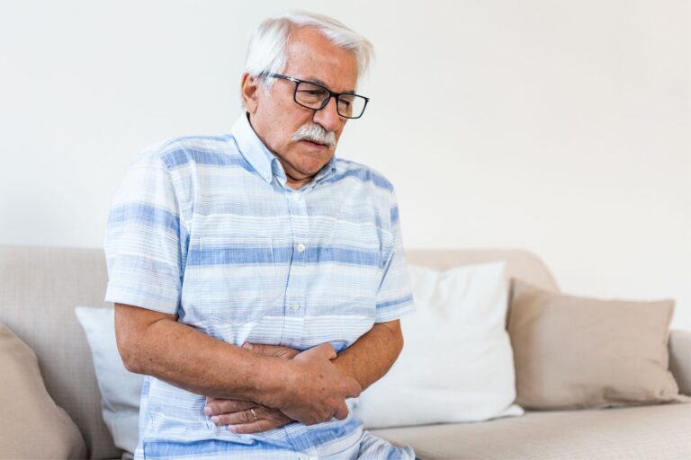 This is a photo of a man sitting on a sofa. He looks in pain, and his clutching around his stomach. It is the cover image to the recording of a webinar held by the Urostomy Association on 4 February 2025 on preventing and managing hernias