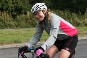 This is a photo of urostomate Julie Mason Wright riding a racing bike. She is wearing a helmet and a pink and grey cycling jacket.