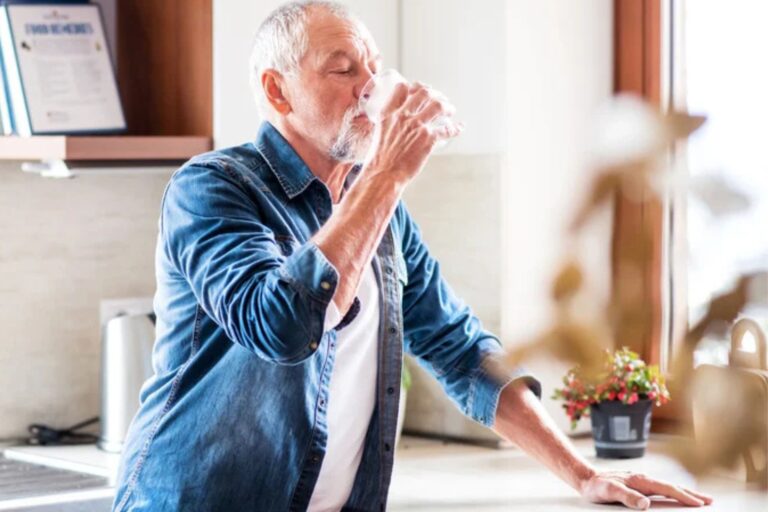 This is a photo of an older man in his kitchen drinking a glass of water. He is wearing a blue shirt over a white t-shirt. He has cropped grey hair and a beard.