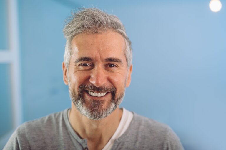 This is a photo of a middle aged man smiling at the camera. He has a beard, greying hair and a grey top. It is the header image for the 'Your neobladder questions answered' page on the Urostomy Association's website