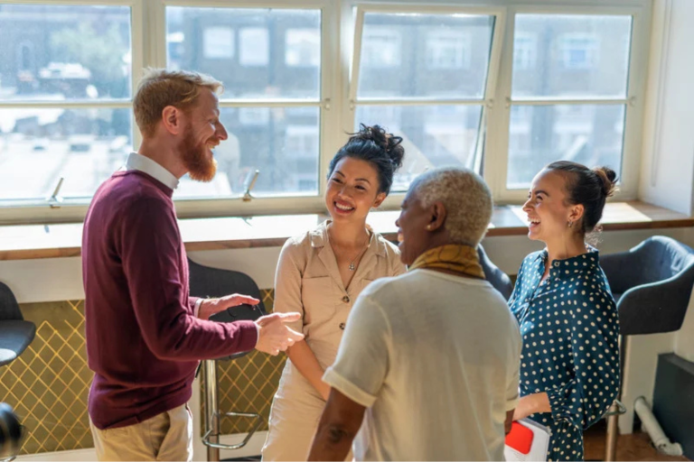 This is a photo of four work colleagues standing in front of a bank of windows laughing. It is the header image for the Urostomy Association's page called 'Confidence and comfort at work'