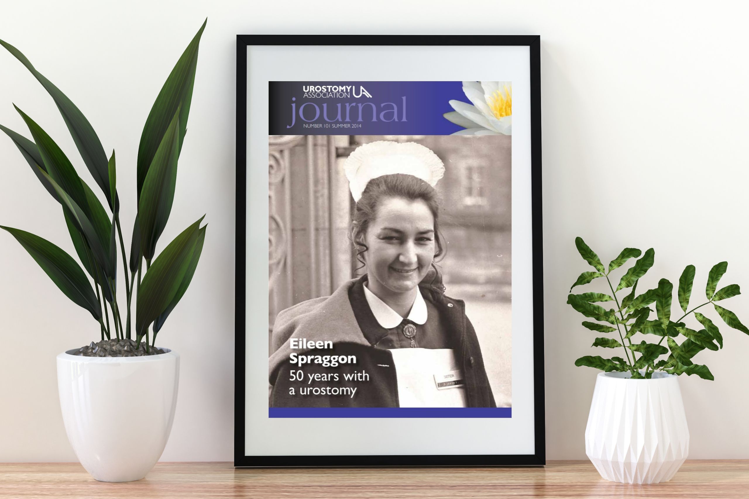 This photo is of a picture frame sitting on a wooden table. In the frame is the cover of Summer14's edition of the Urostomy Association Journal, featuring Eileen Spraggon.