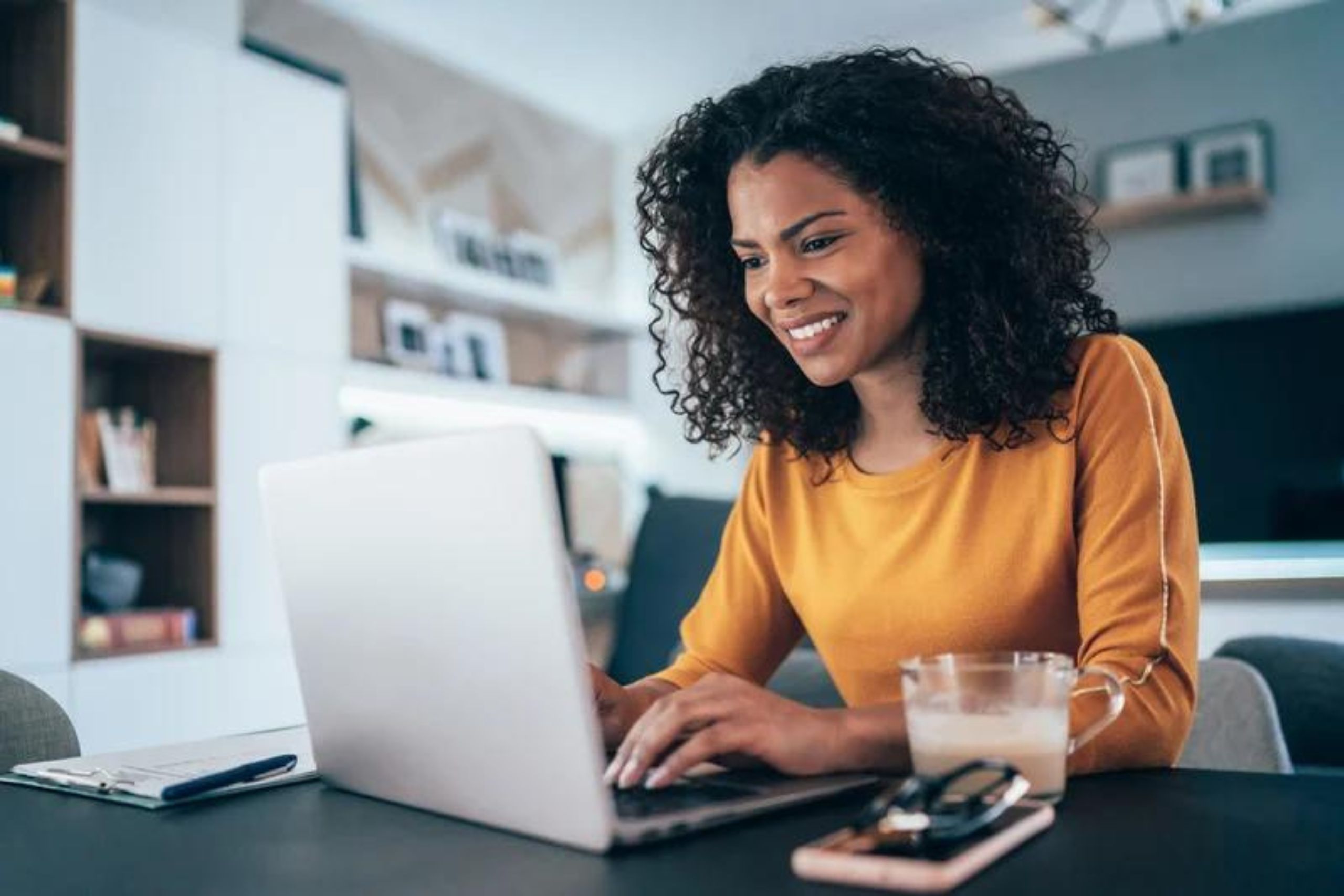 This photo is of a young woman in a mustard-coloured jumper sitting at a table looking at her laptop. She has a cup of coffee, phone, sunglasses and clipboard next to her