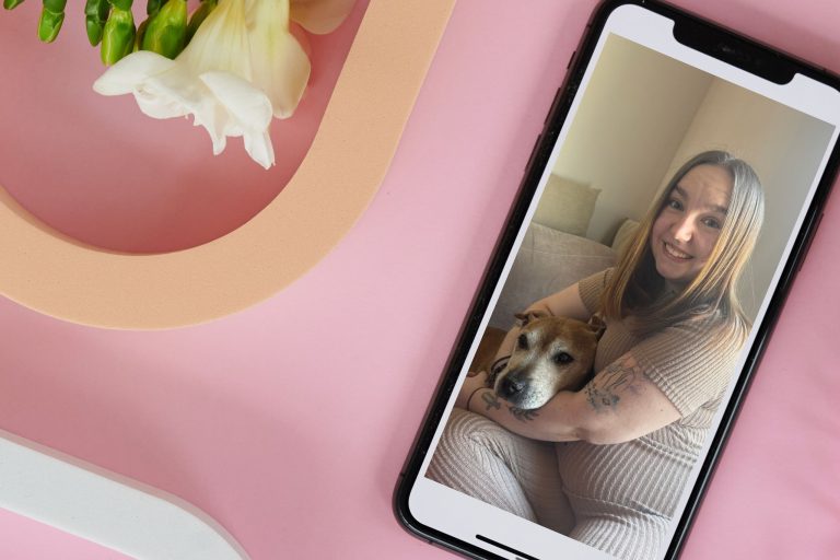 This photo is of a young woman called Aimee. She is sitting with her dog. Their picture has been superimposed over the image of a smart phone. The back ground is pink with a freesia in the top left hand corner.