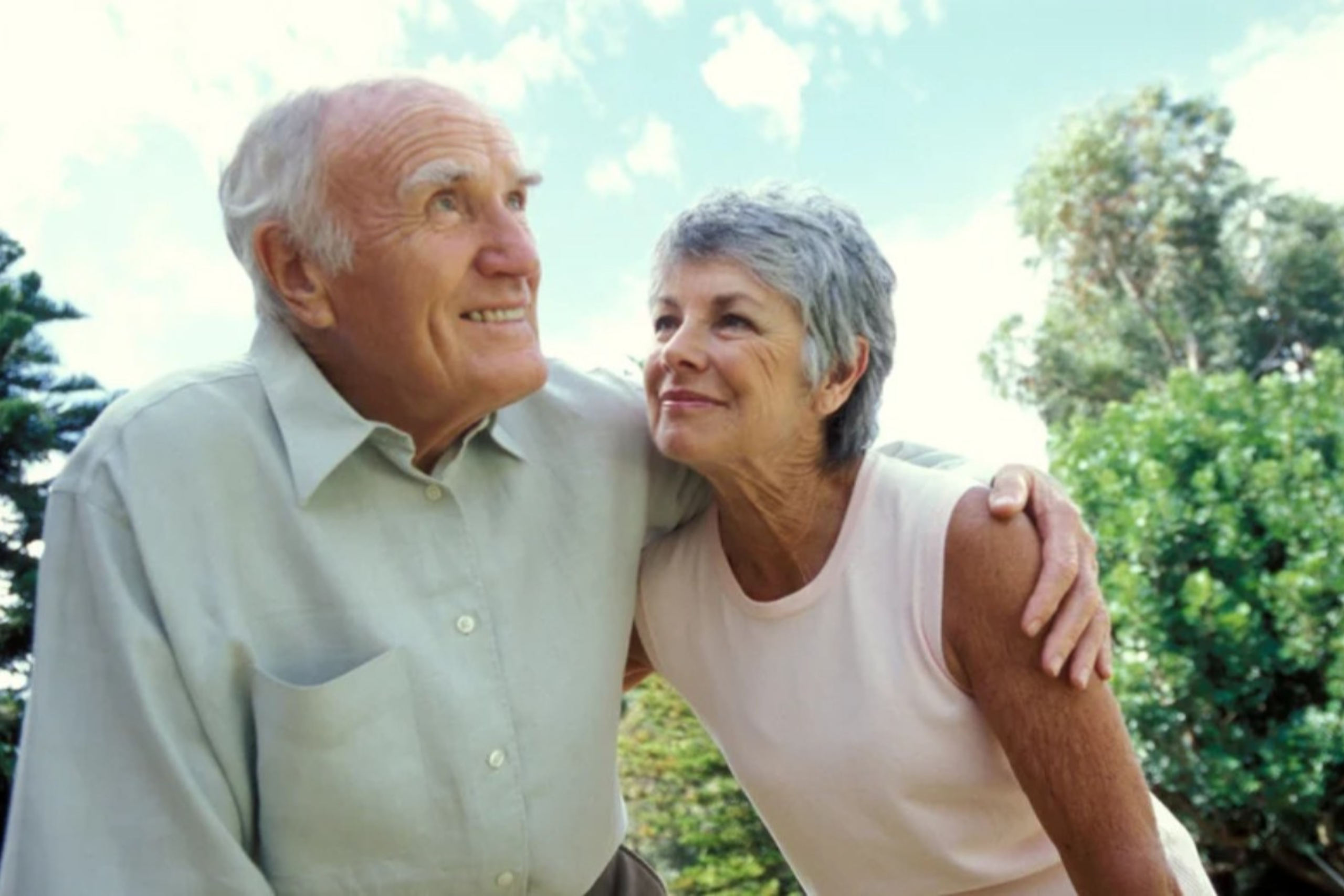 This is a photo of a smiling older couple outdoors. He is looking to the distance and the woman is looking into his face.