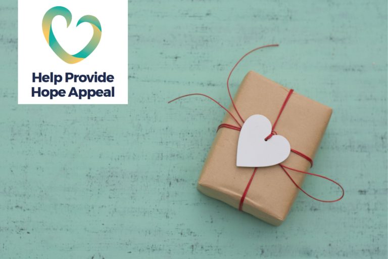 This photo shows a small gift wrapped in brown paper and tied up with red string. Thee is a heart-shaped label on the front. It is resting on a green 'distressed-look' background. To right is a logo with a stylised heart and the words 'Help Provide Hope Appeal' written underneath