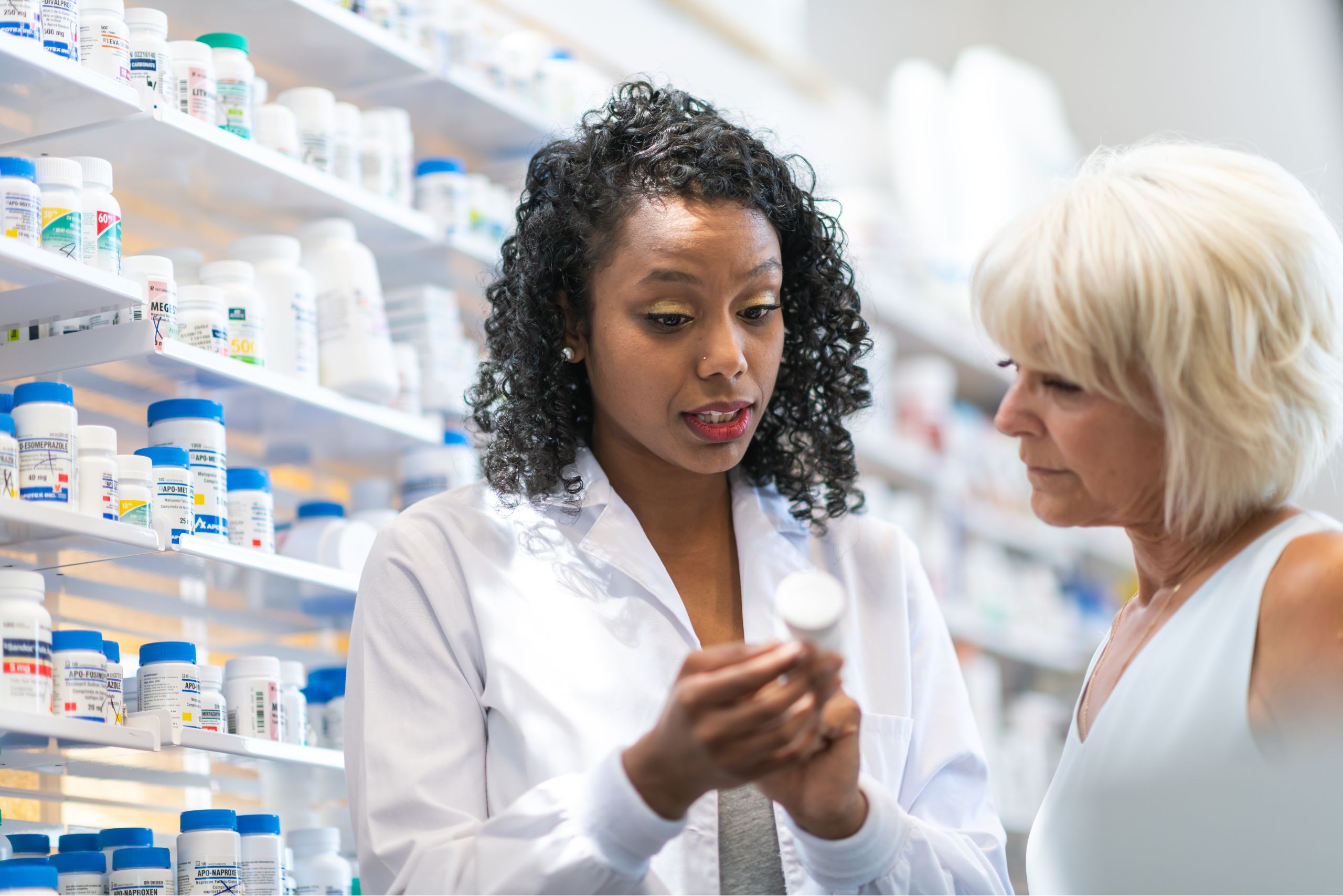 This is a photo set in a pharmacy. A young pharmacist is explaining the labelling on a pill bottle to a customer