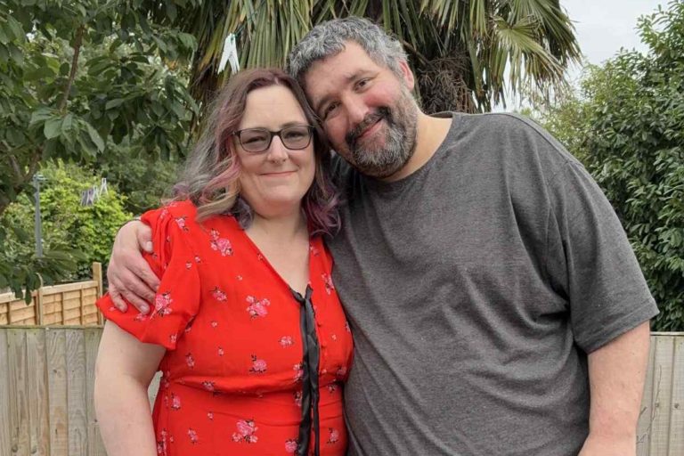 This is a photo of urostomate, Dani, with her partner, Gareth. She is wearing a red v-necked dress. He is wearing a grey t-shirt. He has his arm around her shoulder and they are smiling to the camera.