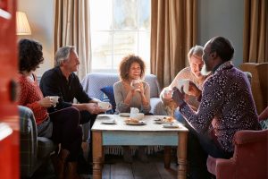 This photo is of a group of five friends sitting around a coffee table laid out with pastries and hot drinks. They're laughing together.