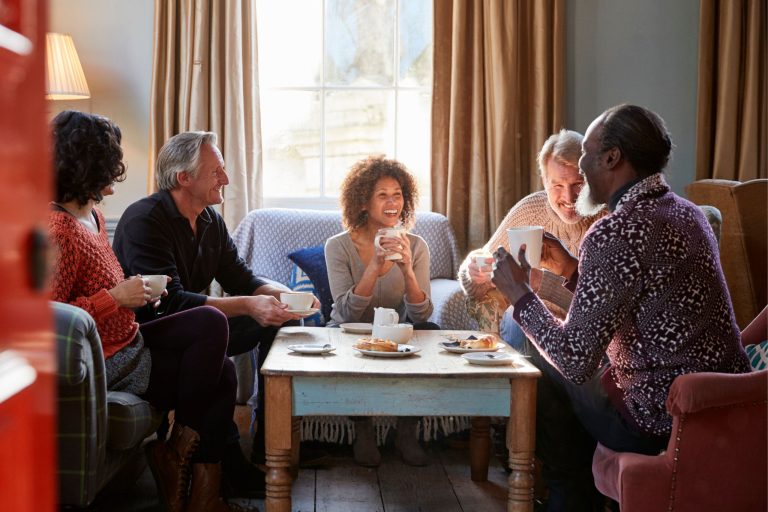 This photo is of a group of five friends sitting around a coffee table laid out with pastries and hot drinks. They're laughing together.