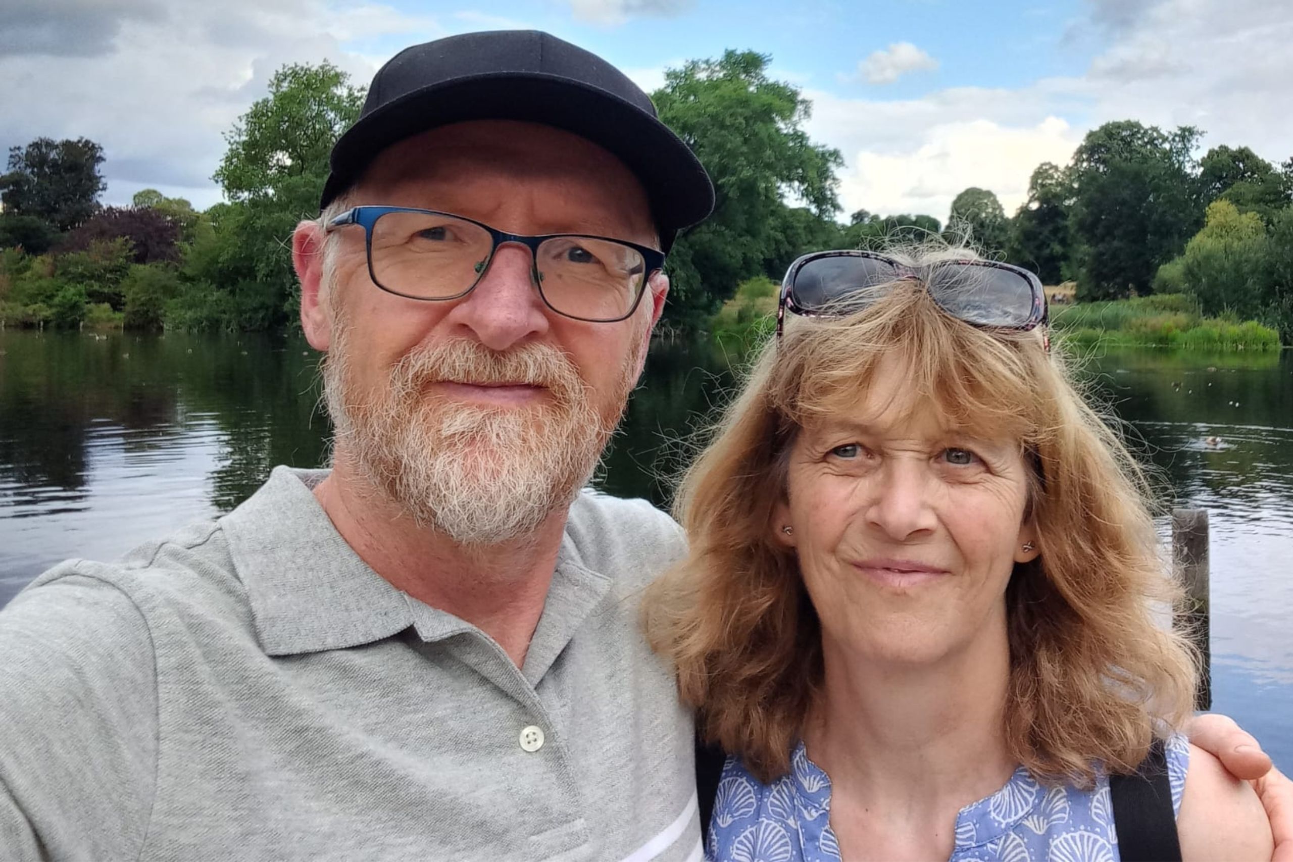 This selfie is of Nicky Rogers and her husband, Neil. Neil has his arm around Nicky, and they are standing in front of a lake or river. The weather looks warm, with a lightly clouded sky.