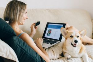 This photo is of a woman sitting on her sofa, with a laptop open on a shopping page. She has a payment card in her left, and is stroking a dog with her right.