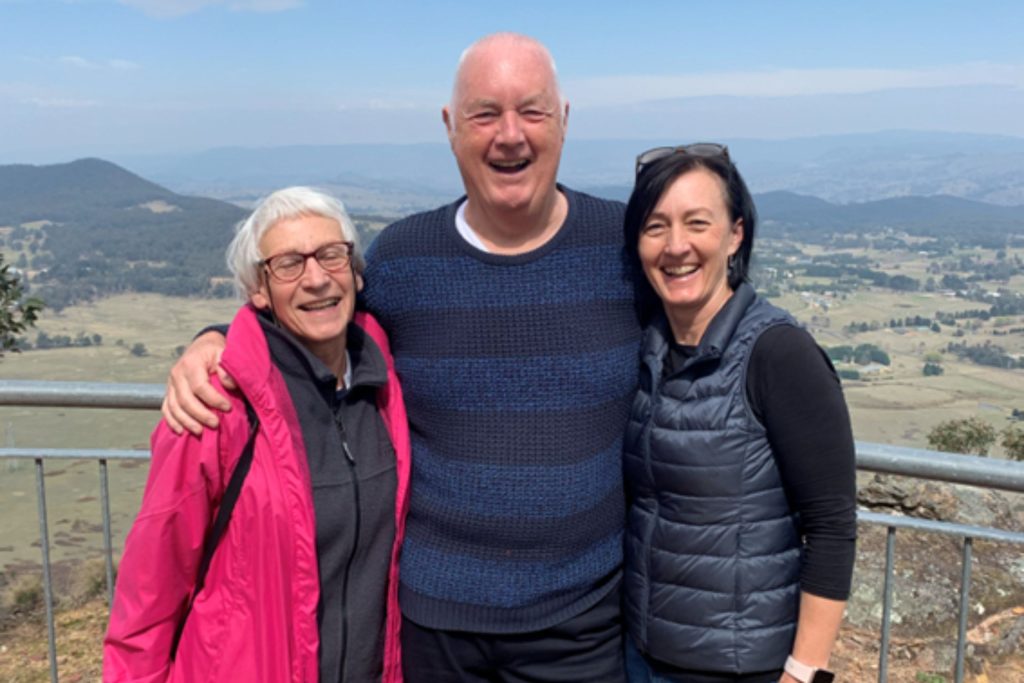 This photo is of Tony and  family. They are standing on a hill and the sky is blue behind them. They are smiling at the camera, and Tony is in the middle with him arms around the women on either side of him. 