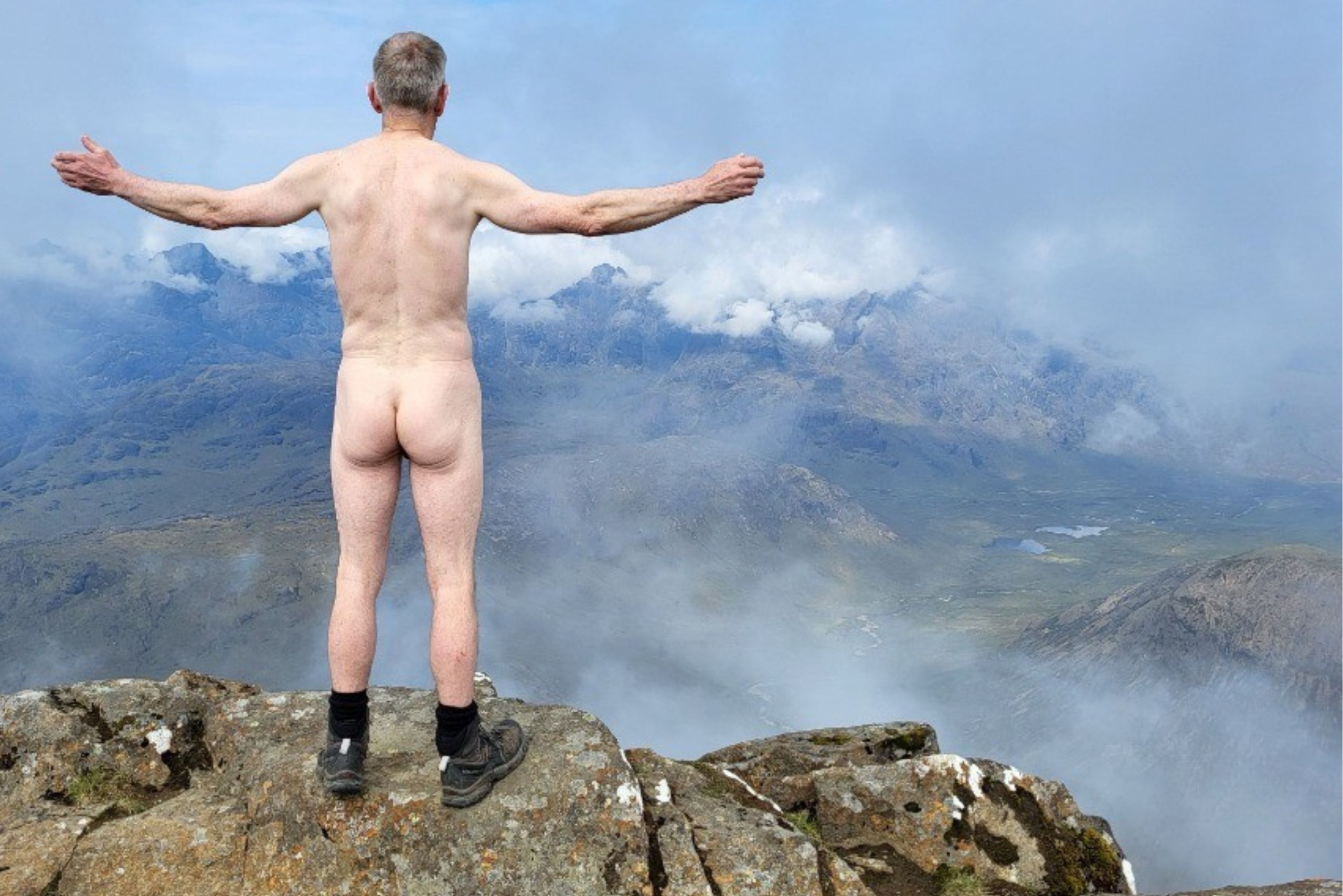 This photo shows Billy standing on a mountain. He is naked apart from his boots. He is facing away from the camera with his arms outstretched.