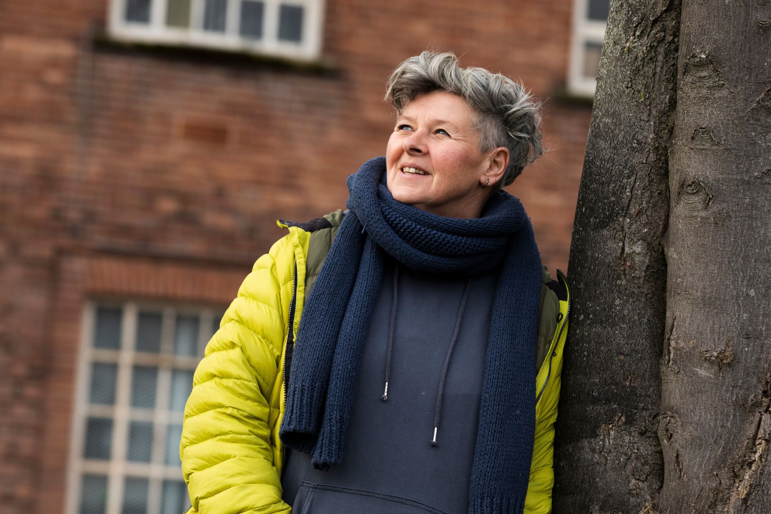 This photo shows a woman leaning against a tree, looking upward thoughtfully. She is wearing a lime green jacket and a great sweatshirt and scarf.