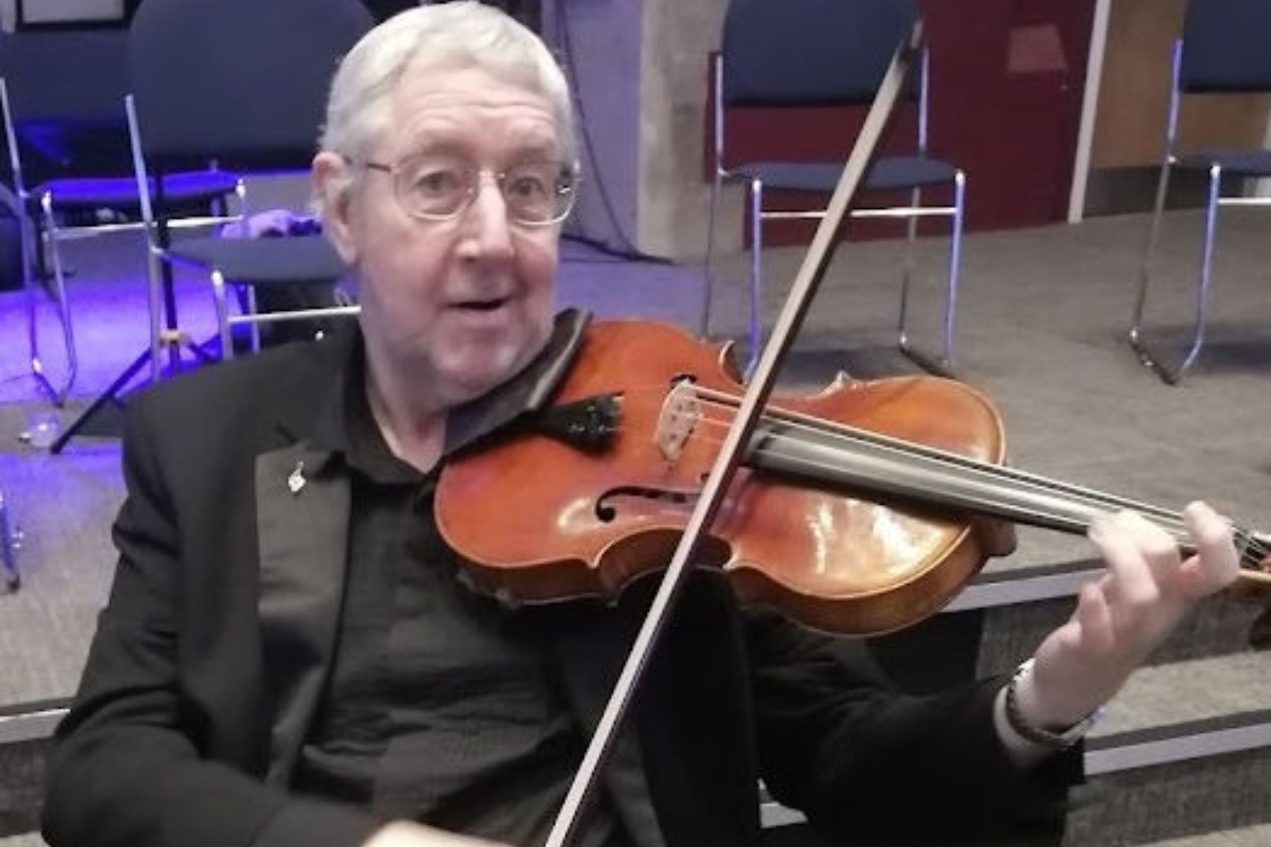 In this photo of Peter he is looking straight at the camera. He is wearing glasses, a black shirt and is playing a violin.