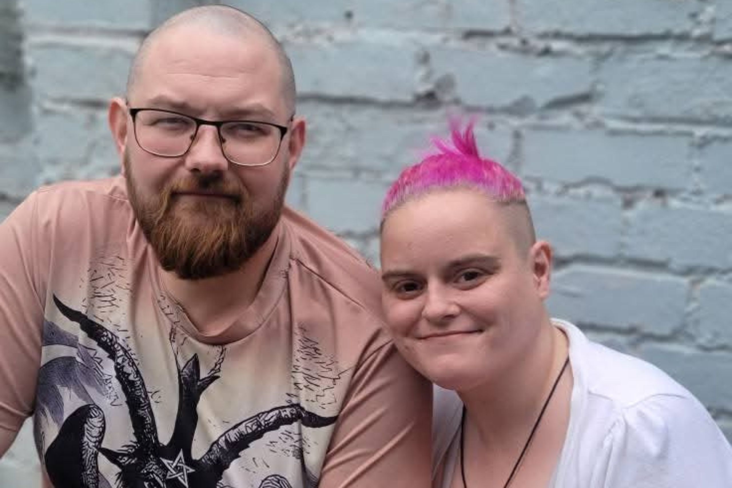 This is a head and shoulders shot of a young couple. Tia, on the right, has short pink hair. Stephen has a beard and glasses. They are smiling at the camera.