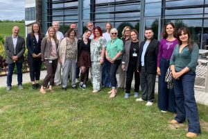 This photo shows 16 people in a group shot. They are members of the organisation, Invisible. They are outdoors in front of a Coloplast building in Copenhagen.