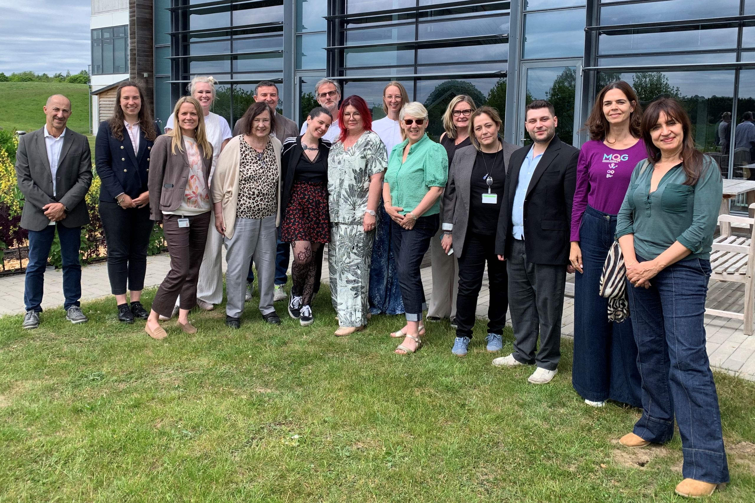This photo shows 16 people in a group shot. They are members of the organisation, Invisible. They are outdoors in front of a Coloplast building in Copenhagen.