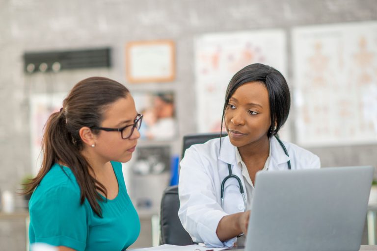 This photo is of two women in a consultation. The clinician is seated on the right in a white medical coat, and she is pointing to something on a laptop to the patient on the left.