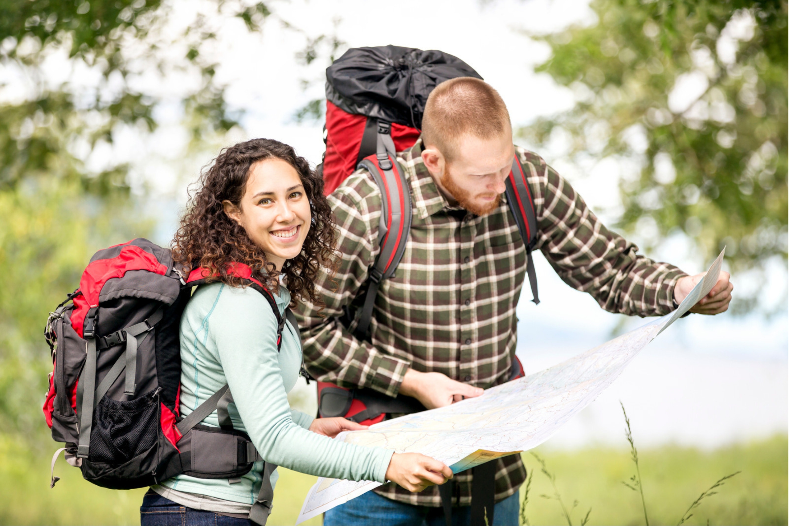 This photo shows a young couple outdoors, with back packs. He is looking at a map, and she is smiling at the camera