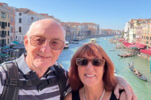 This photo shows Hugh Rogers and his wife Tina on a bridge in Venice. They are taking a selfie, and smiling